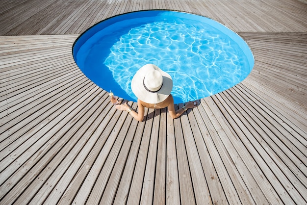 Vrouw in grote zonnehoed met cocktaildrank ontspannen bij het ronde zwembad met blauw water buitenshuis. Bovenaanzicht