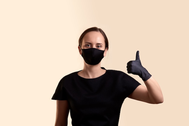 Vrouw in het zwarte masker en de handschoenen die van het kledingsgezicht vthumb en over beige muur tonen