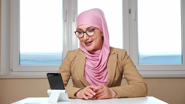 Vrouw in hijab en bril zit aan een tafel en praat via video-oproep aan de telefoon