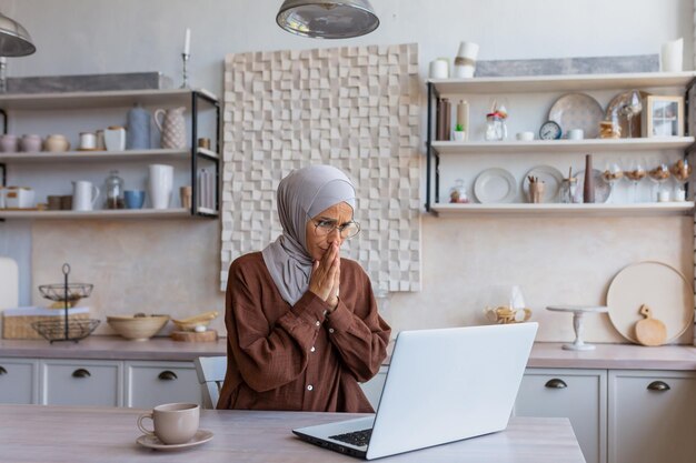 Vrouw in hijab thuis overstuur en teleurgesteld leesbericht van moslimvrouw op laptopscherm