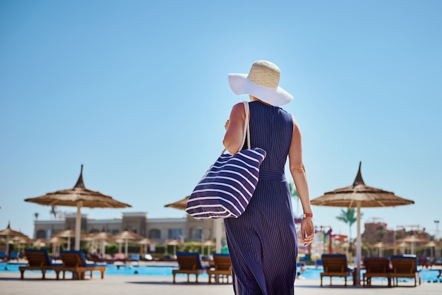 Vrouw in hoed en blauwe jurk gaan voor een wandeling in het hotel resort bij zwembad met strandtas