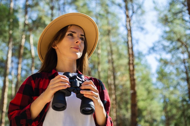 Foto vrouw in hoed en rood geruit overhemd met verrekijker in het bos.