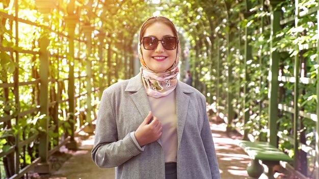 Vrouw in hoofddoek en zonnebril glimlachend in tuinboog, zonlicht