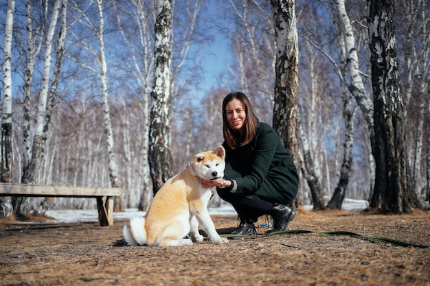 Vrouw in jas en een akita inu-puppy in een winterberkenpark kijken naar de camera