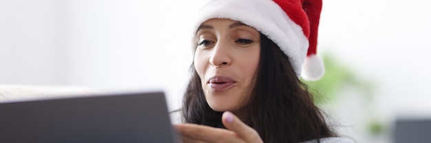 Vrouw in kerstman hoed glimlacht en blaast kus op laptopcomputer