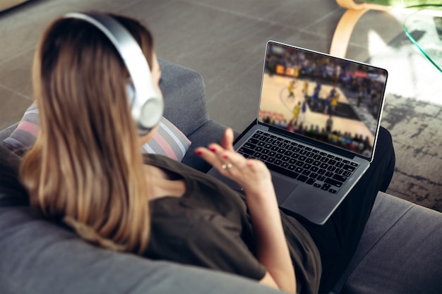 Vrouw in koptelefoon met laptop kijken naar sport, basketbal online streaming van kampioenschap. Nieuwe regels tijdens de uitbraak van het coronavirus en de afsluiting van landen. Concept van sport, competitie, technologieën.