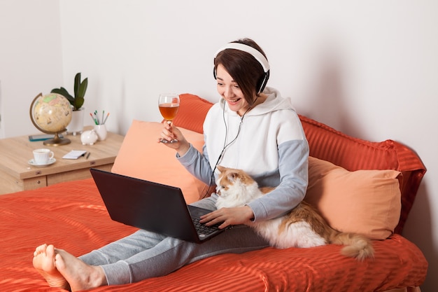 Vrouw in koptelefoon zittend op het bed met een kat. Thuis online onderwijs en werk in comfortabele en comfortabele omstandigheden.