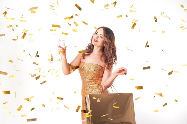 Vrouw in mode gouden kleding met een gouden papieren zak en een glas mousserende wijn en gouden confetti vieren op feestje, witte studio achtergrond