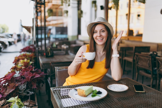 Vrouw in openlucht straat coffeeshop café zittend aan tafel in gele kleding hoed met kopje cappuccino, cake, ontspannen in restaurant tijdens vrije tijd