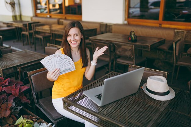 Vrouw in openlucht straat coffeeshop café zittend met moderne laptop pc-computer, houdt in de hand een stel dollars bankbiljetten