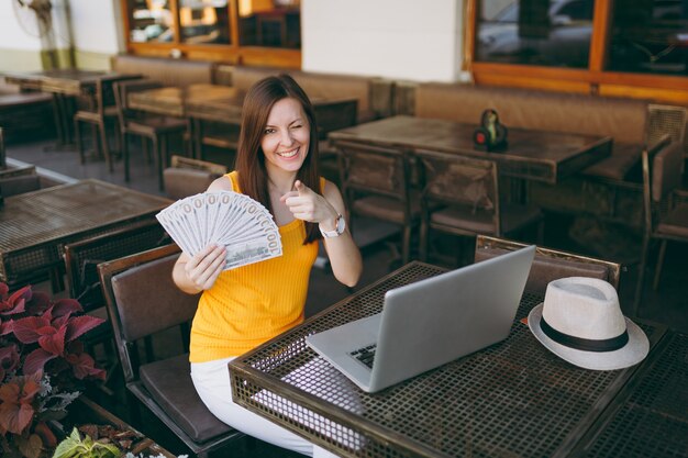 Vrouw in openlucht straat coffeeshop café zittend met moderne laptop pc-computer, houdt in de hand een stel dollars bankbiljetten