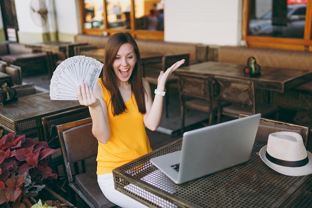 Vrouw in openlucht straat coffeeshop café zittend met moderne laptop pc-computer, houdt in de hand een stel dollars bankbiljetten