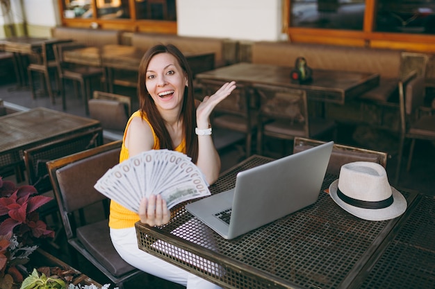 Vrouw in openlucht straat coffeeshop café zittend met moderne laptop pc-computer, houdt in de hand een stel dollars bankbiljetten