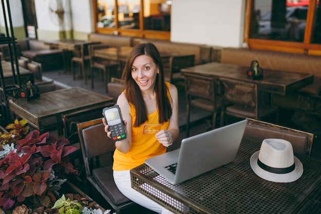 Vrouw in openlucht straat coffeeshop zittend met laptop pc-computer, houdt draadloze moderne bankbetaalterminal om creditcardbetalingen te verwerken
