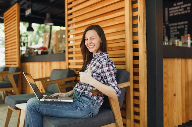 Vrouw in openlucht straat zomer coffeeshop houten café zittend in vrijetijdskleding, werken op moderne laptop pc-computer