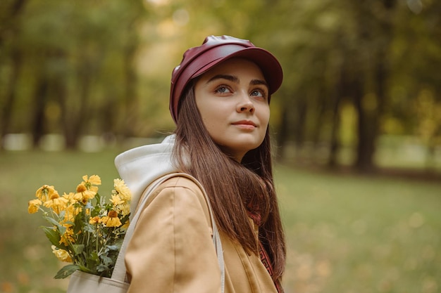 vrouw in pet en greppel met boeket gele bloemen in zak in herfstpark