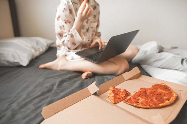 Vrouw in pyjama werkt op een laptop in bed en eet een pizzabezorger.