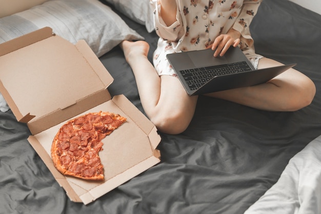 Vrouw in pyjama zit op bed met een doos pizza en maakt gebruik van een laptop.