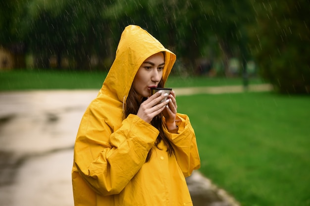 Vrouw in regenjas drinkt hete thee in zomerpark, regenachtige dag. Alleen vrouwelijke persoon in regenkaap op wandelpad, nat weer in steegje