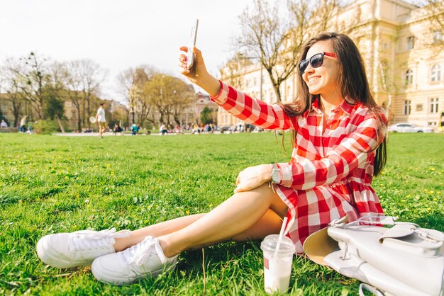 Foto vrouw in rode jurk zit in park op groen gras met smartphone