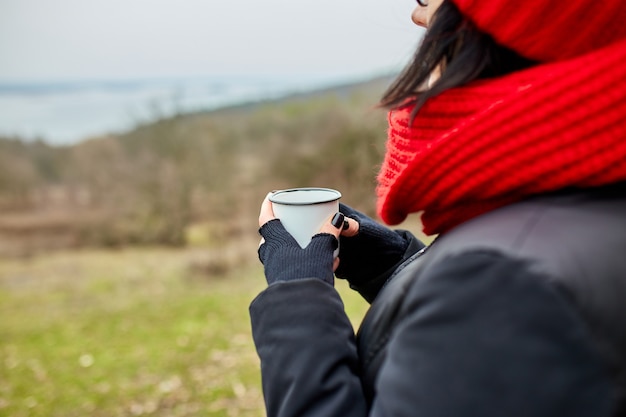 Vrouw in rode muts en sjaal, thee drinken uit metalen beker buiten, Travel concept, herfst achtergrond landschap.