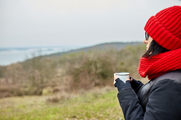 Vrouw in rode muts en sjaal, thee drinken uit metalen beker buiten, travel concept, herfst achtergrond landschap.