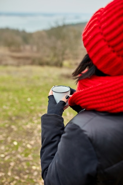 Vrouw in rode muts en sjaal, thee drinken uit metalen beker buiten, Travel concept, herfst achtergrond landschap.