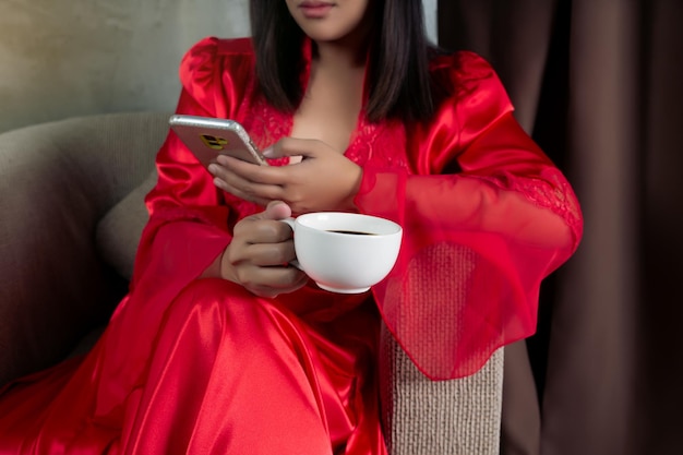 Vrouw in rode nachtjapon die een koffiekopje vasthoudt en naar een smartphone kijkt