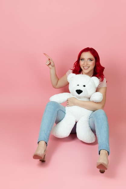 Vrouw in spijkerbroek met rood haar houdt een grote witte teddybeer vast en wijst met haar wijsvinger