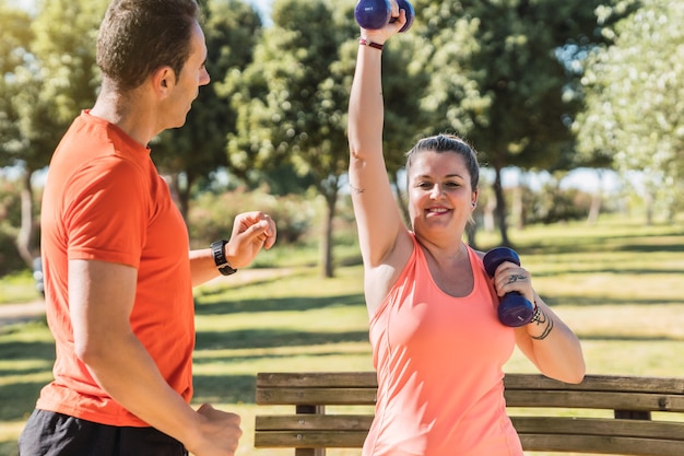 Vrouw in sportkleding die gewichten doet met een personal trainer naast haar in een park.