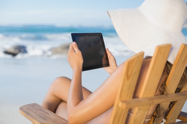 Vrouw in strohoed het ontspannen in ligstoel op het strand die tabletpc met behulp van