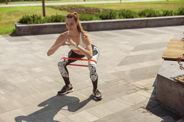 Vrouw in trainingspak doet squats met elastische buiging in stadspark