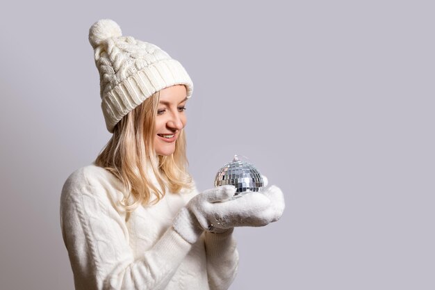 Foto vrouw in trui een pet met een bubo en handschoenen glimlacht en kijkt naar een kerstboom speelgoed disco bal