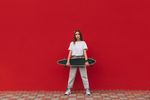 vrouw in vrijetijdskleding op straat poseren voor de camera met een longboard in handen op een rode muurachtergrond