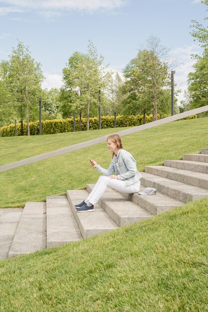 Vrouw in vrijetijdskleding sms't op haar mobiele telefoon zittend op de trap in het park