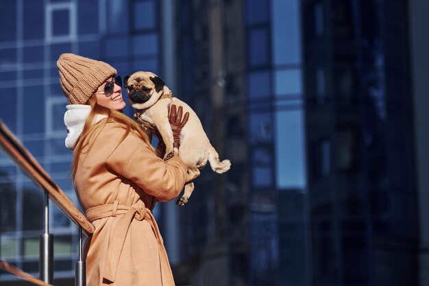 Vrouw in warme kleren heeft haar kleine mopshond op handen in de buurt van een bedrijfsgebouw dat op de achtergrond staat.