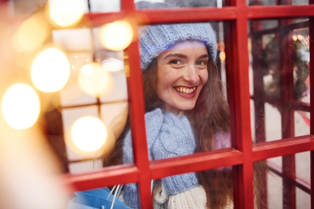 Vrouw in warme kleren veel plezier in het telefoonstation. Kijk door het glas.