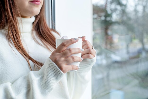 Vrouw in warme witte wintertrui die thuis op de vensterbank zit