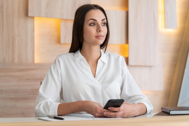 Vrouw in wit overhemd zittend aan tafel met telefoon in handen en notebook met laptop op tafel