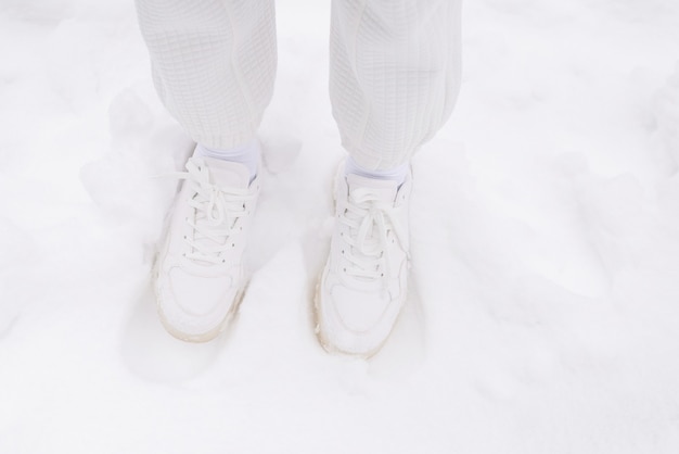 Vrouw in witte broek en witte sneakers staat in een sneeuw.