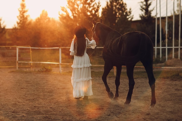 Vrouw in witte jurk en zwarte hoed met haar paard bij zonsondergang buiten op de ranch