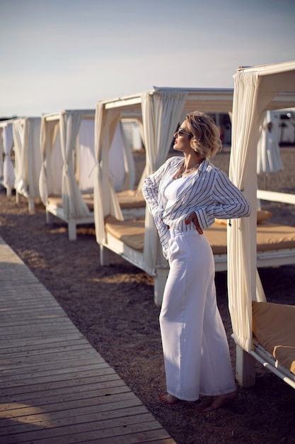 Vrouw in witte kleren en zonnebril staat op het strand in de buurt van houten