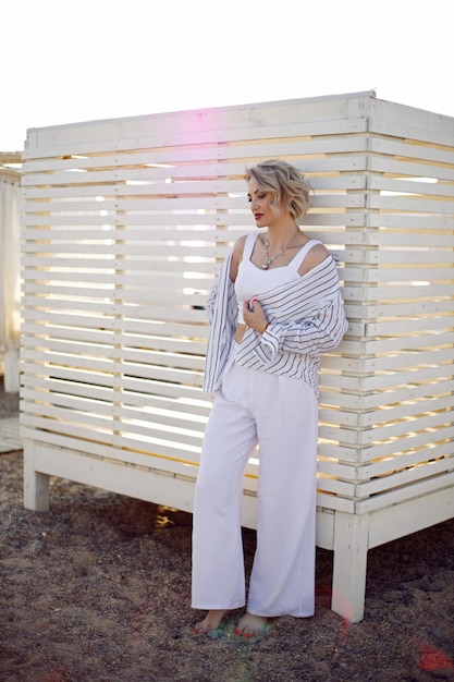 Vrouw in witte kleren en zonnebril staat op het strand in de buurt van houten