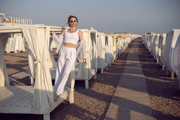 Vrouw in witte kleren en zonnebril staat op het strand in de buurt van houten