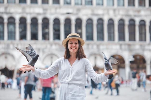 Vrouw in witte kleren met strohoed die plezier heeft met duiven op het plein Piazza San Marco . van Venetië