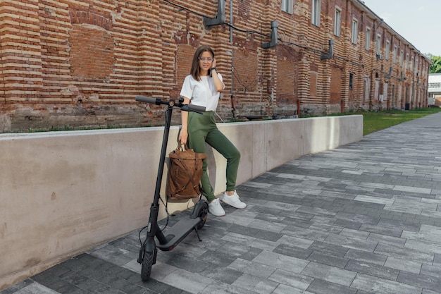 Vrouw in witte sneakers en leren rugzak staande in de buurt van elektrische scooter