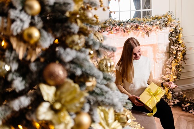 Vrouw in witte trui, warme en gezellige avond in een kerstinterieur, kerstboom versierd met lichtjes, slinger die binnenshuis oplicht. Zachte focus