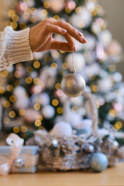 Vrouw in witte warme wollen trui met speelgoed decoratieve bal in handen op de achtergrond van een kerstboom. Kerstmis, nieuwjaarsconcept voor vakantieviering. Selectieve focus