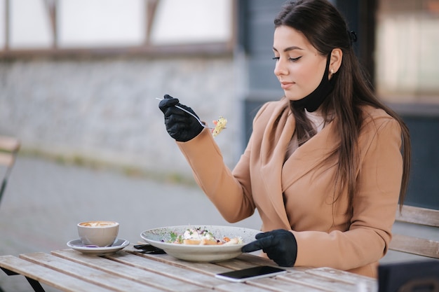 Vrouw in zwart gezichtsmasker zittend in café en voorbereid op het eten van veganistische salade.