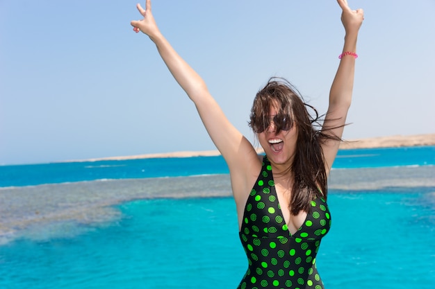 Vrouw in zwart-groen zwempak die haar handen opsteekt en lacht op het jacht op een zonnige zomerdag, prachtige turquoise zee op de achtergrond